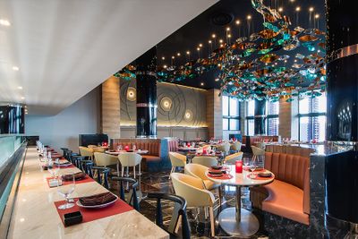 Modern restaurant interior with elegant decor, colorful ceiling art, round tables, cushioned chairs, and large windows offering city views.