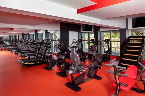 A modern gym with red flooring, featuring treadmills, stationary bikes, and step machines, lined up in front of large windows.