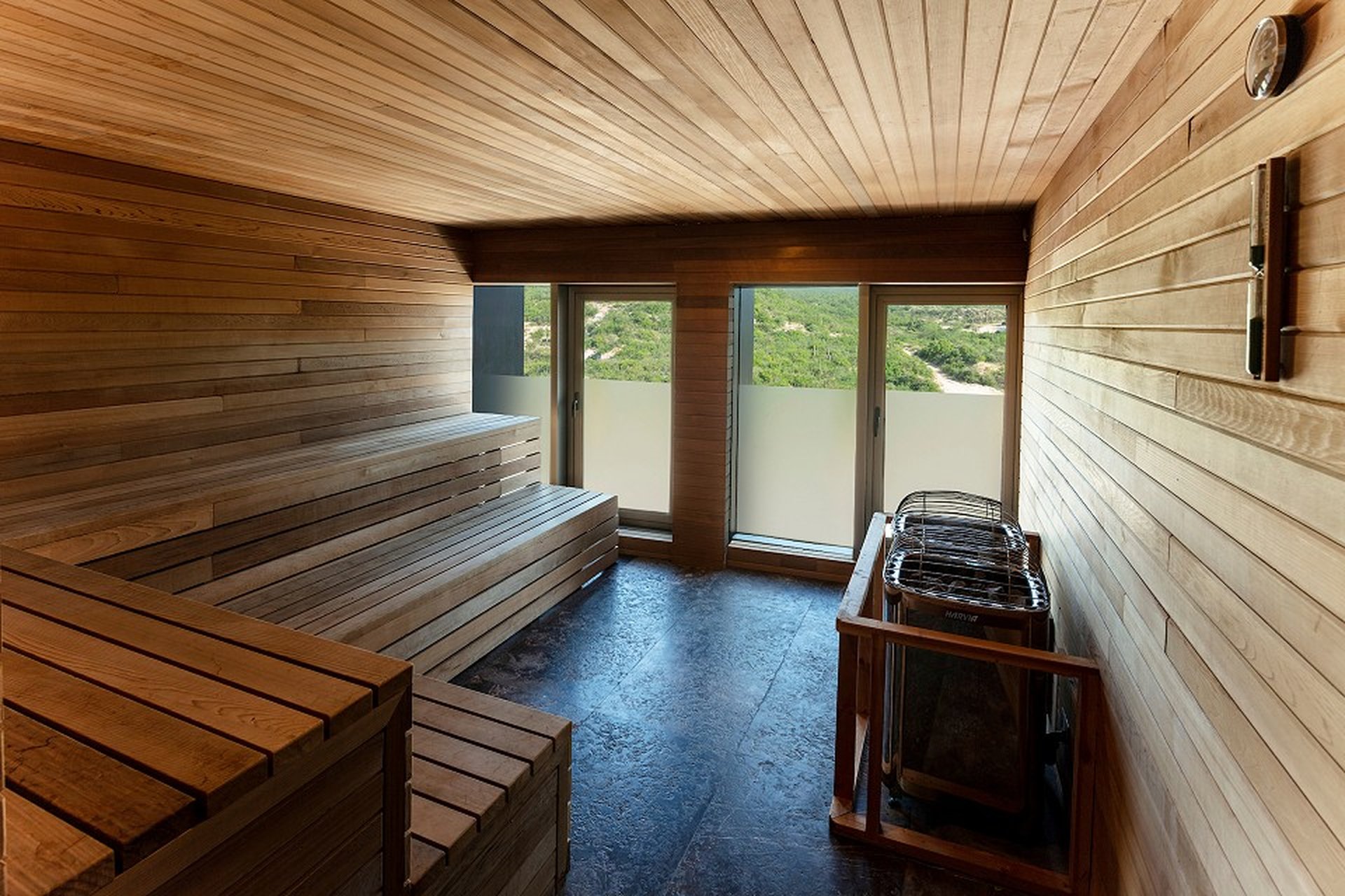 Wooden sauna interior with benches, a heater, and large windows overlooking a green landscape outside.