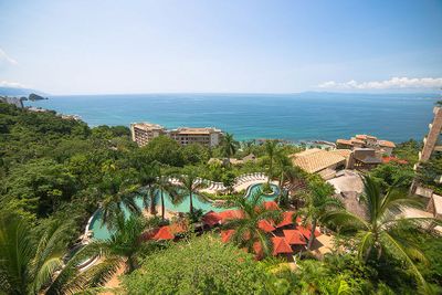Aerial view of a coastal resort with a pool, surrounded by lush greenery and overlooking a vast, calm ocean under a clear blue sky.
