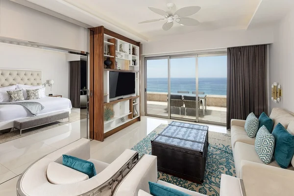 Luxurious hotel suite with ocean view, featuring a living area, large bed, TV, elegant furniture, and a balcony with glass doors.
