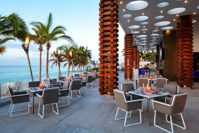 Outdoor restaurant with modern decor, featuring gray chairs, set tables with red candles, palm trees, and ocean view under a blue sky.