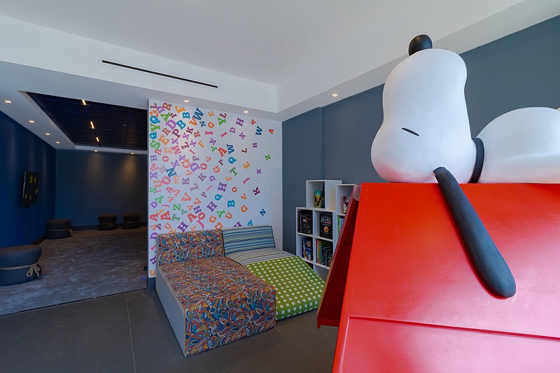Children's playroom with Snoopy statue on a red doghouse, colorful alphabet wall, patterned bed, and blue walls with seating area.