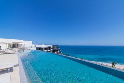 Infinity pool overlooking a clear blue ocean, with white buildings and a cloudless sky in the background.