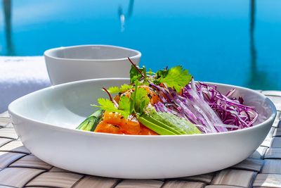 A white bowl with sliced salmon, avocado, cucumber, and shredded purple cabbage, garnished with cilantro, placed on a woven mat by a pool.