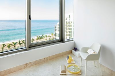 Bright room with ocean view, large window, white chair, and spa items on the floor. Palm trees and beach visible outside.