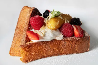 A slice of French toast topped with whipped cream, raspberries, strawberries, blackberries, banana slices, and almond flakes.