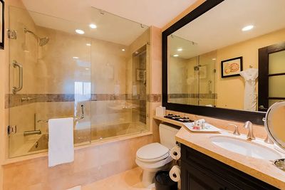 Modern bathroom with a glass-enclosed bathtub, large mirror, toilet, and sink on a marble countertop. Soft lighting and beige tones.