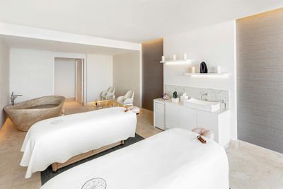 Bright spa room with two massage tables, a stone bathtub, white chairs, and a sink. Minimalist decor with neutral tones and soft lighting.
