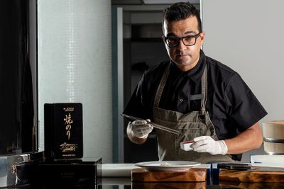 Chef in a black shirt and apron, holding chopsticks, prepares a dish in a modern kitchen with a box of ingredients nearby.