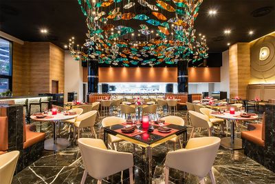 Elegant restaurant interior with modern decor, featuring colorful ceiling art, round tables with red candles, and beige chairs on marble flooring.