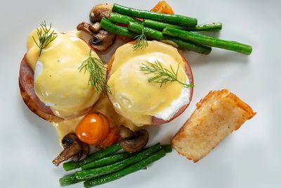 Eggs Benedict with hollandaise sauce, green beans, mushrooms, cherry tomatoes, and a hash brown on a white plate, garnished with dill.