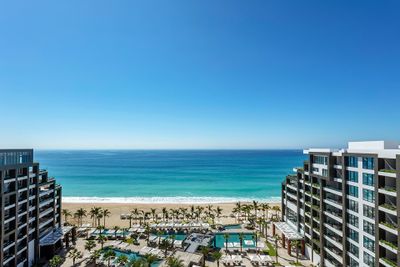 Aerial view of a beachfront resort with modern buildings, palm trees, pools, and lounge areas overlooking a clear blue ocean under a bright sky.
