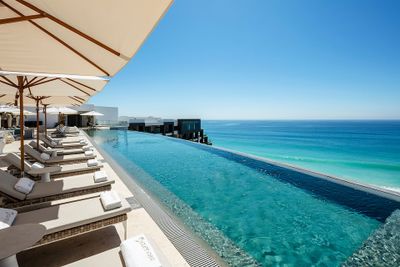 Infinity pool overlooking a clear blue ocean, with sun loungers and umbrellas lining the poolside under a bright, cloudless sky.