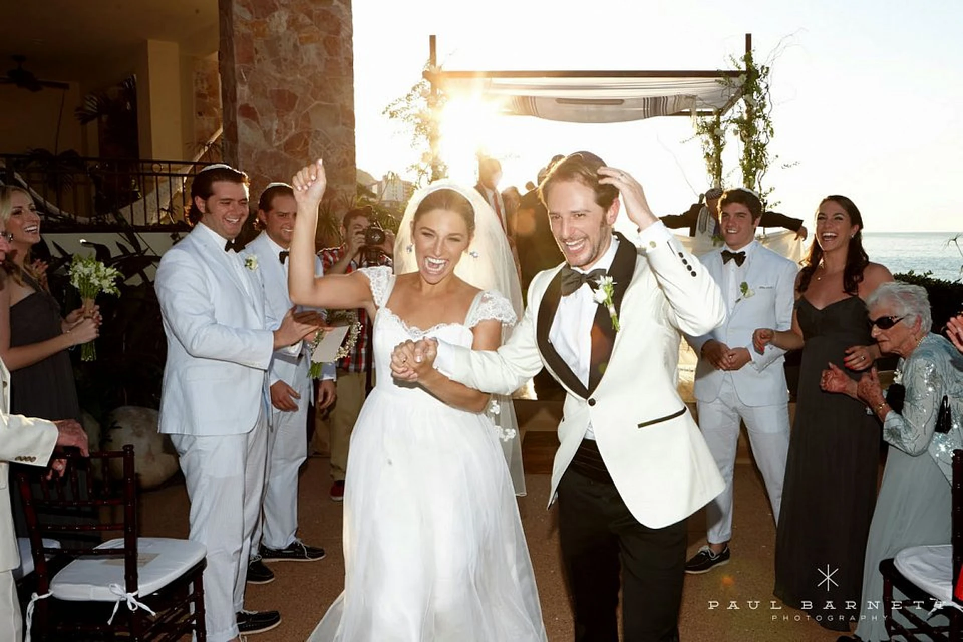 Bride and groom joyfully walk down the aisle, surrounded by cheering guests, with the sun setting in the background.