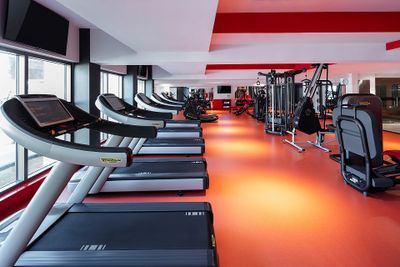 Modern gym with treadmills, weight machines, and bright red flooring. Large windows allow natural light to illuminate the space.