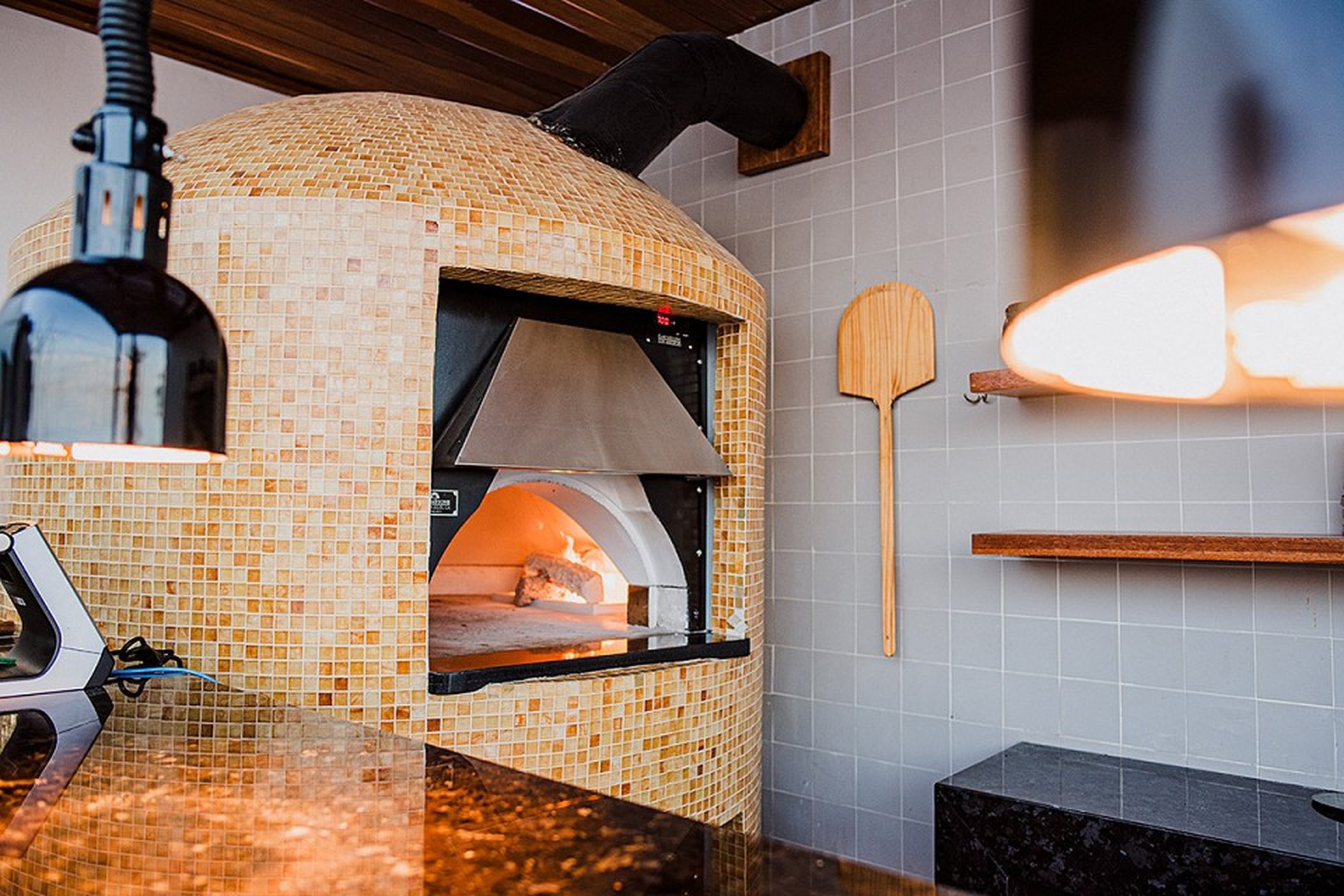 A tiled wood-fired pizza oven with a lit fire inside, a pizza paddle hanging on the wall, and a black countertop in the foreground.