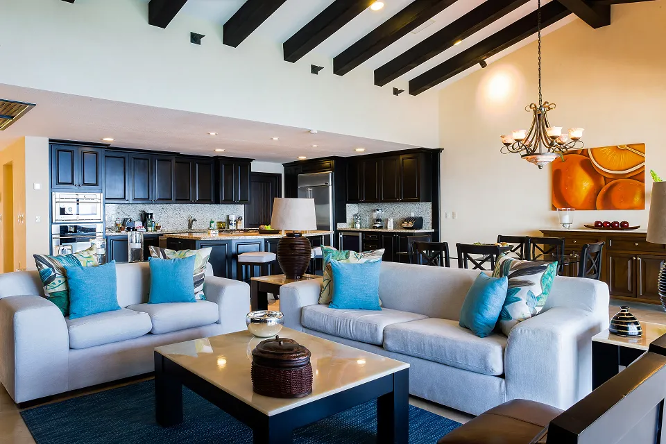 Luxurious living area with elegant sofas, vibrant cushions, dark wood kitchen, and dining space under a chandelier in a high-ceilinged room.