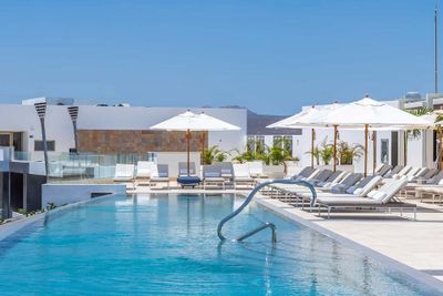 Luxurious outdoor pool with white lounge chairs and umbrellas, surrounded by modern white buildings under a clear blue sky.