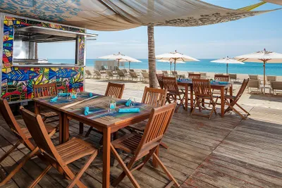 Beachside restaurant with wooden tables and chairs under a canopy, overlooking the ocean. Umbrellas and lounge chairs are on the sandy beach.