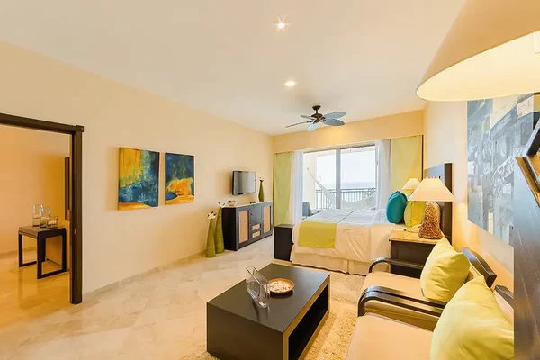 Spacious hotel room with a bed, sofa, and TV. Large window with curtains opens to a balcony. Warm lighting and abstract wall art enhance the decor.