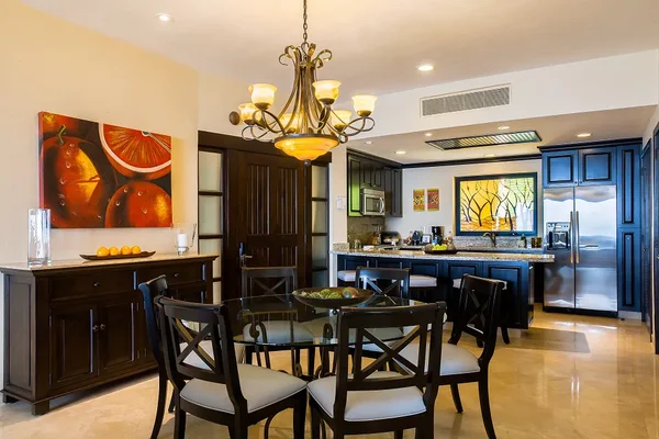 Elegant kitchen and dining area with a glass table, dark wood furniture, colorful fruit artwork, and a modern chandelier.