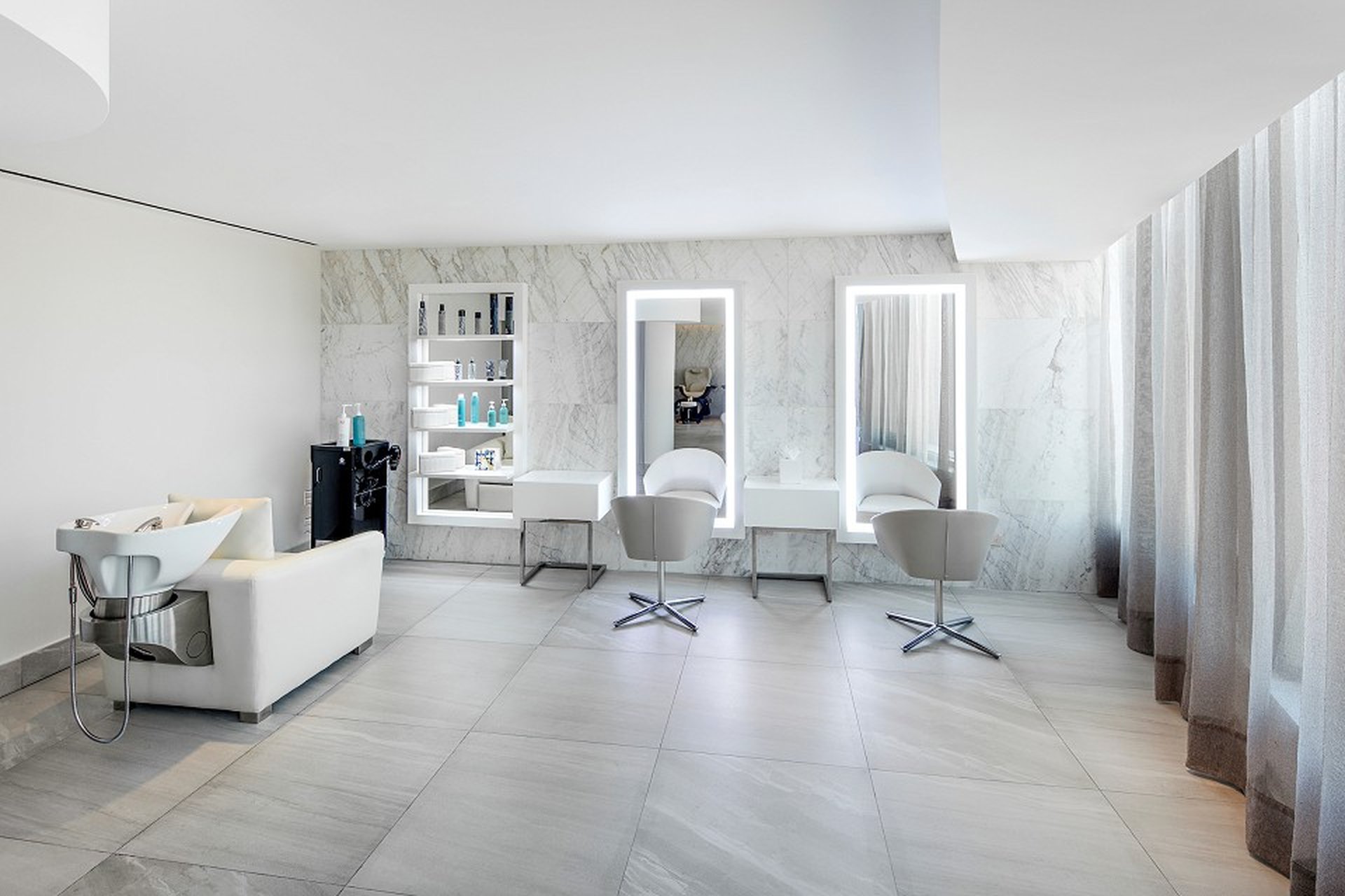 Modern salon interior with marble walls, two styling stations with illuminated mirrors, white chairs, and a shampoo station on the left.