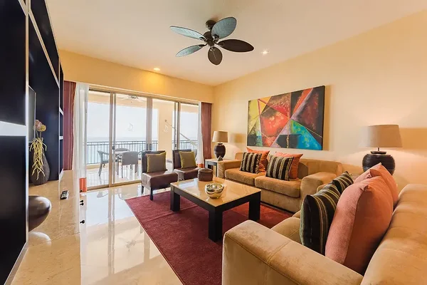 Modern living room with beige sofas, colorful cushions, abstract wall art, ceiling fan, and sliding glass doors leading to a balcony.