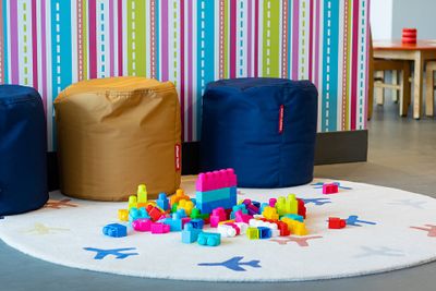 Colorful building blocks on a round rug with airplane designs, surrounded by blue and brown poufs against a striped wall.