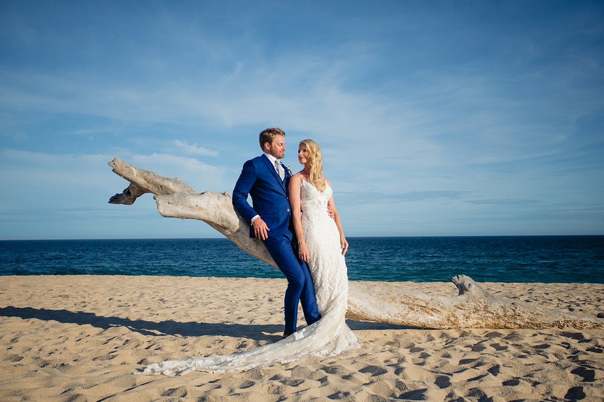 Bride in a white dress and groom in a blue suit pose by driftwood on a sandy beach with a blue ocean backdrop.