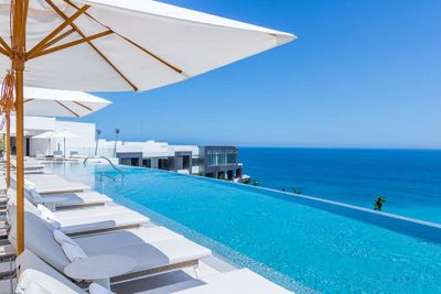 Infinity pool with white lounge chairs and umbrellas overlooking a clear blue ocean under a bright sky. Modern white buildings in the background.