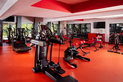 Modern gym with red flooring, featuring treadmills, exercise bikes, and weightlifting equipment. Large windows provide natural light.