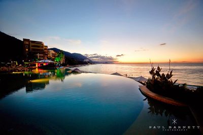 Infinity pool overlooking a serene ocean at sunset, with mountains and buildings in the background, under a colorful sky.
