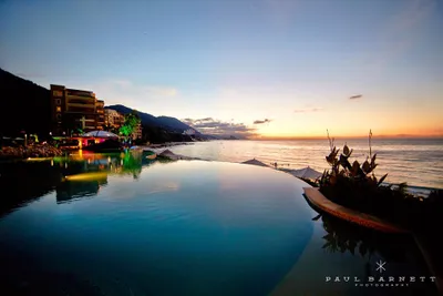 Infinity pool overlooking a serene ocean at sunset, with mountains and buildings in the background, under a colorful sky.