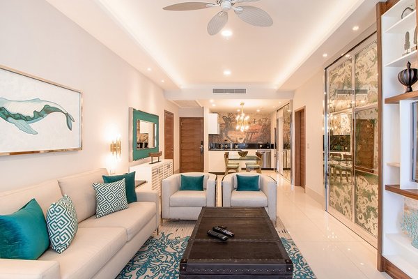 Modern living room with beige sofa, teal accents, and a large mirror. Open layout leads to a kitchen with a chandelier and bar stools.
