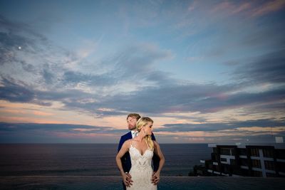 A couple in formal attire stands by the ocean at sunset, with a dramatic sky and modern building in the background.