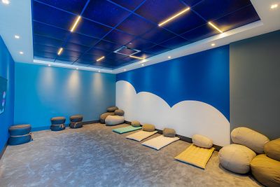 Room with blue walls and ceiling, cloud mural, soft lighting, cushions, and floor mats arranged for relaxation or meditation.