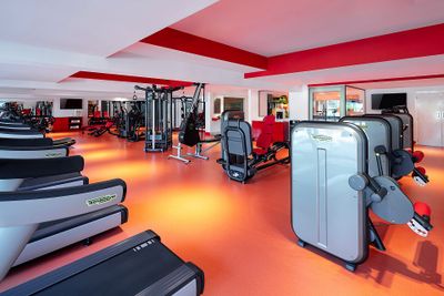 Modern gym with red and white decor, featuring treadmills, weight machines, and exercise equipment on an orange floor.