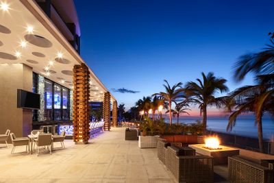 Outdoor patio with modern seating, fire pits, and palm trees at sunset. A bar with blue lighting is on the left, overlooking the ocean.