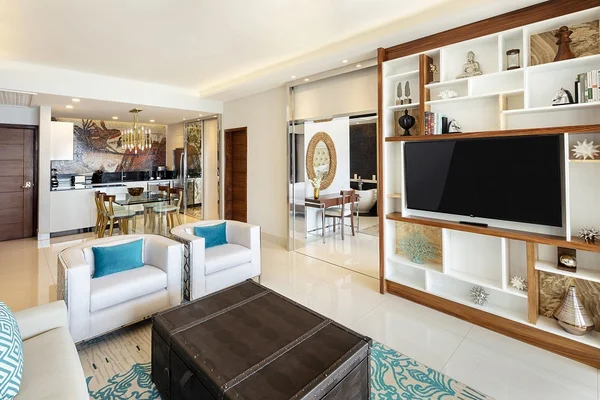 Modern living room with white shelves, a flat-screen TV, two armchairs, and a large trunk-style coffee table. Open kitchen and dining area in the background.