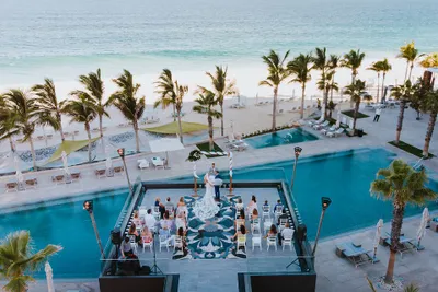 Aerial view of a beachside wedding ceremony by a pool, with guests seated and palm trees lining the sandy shore.