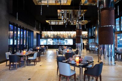 Modern restaurant interior with elegant tables, plush chairs, and ambient lighting. Open kitchen visible in the background.