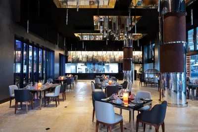 Modern restaurant interior with elegant tables, plush chairs, and ambient lighting. Open kitchen visible in the background.