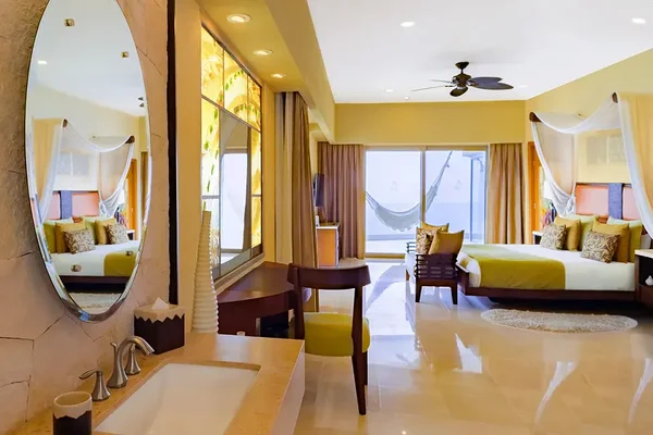 Spacious bedroom with two canopy beds, a hammock by large windows, a round mirror above a sink, and warm yellow accents.
