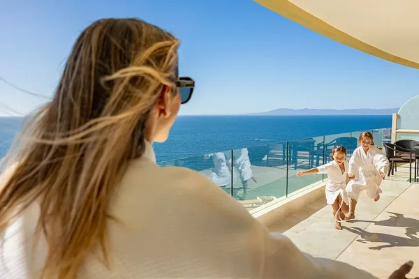 Woman in a robe watches two children joyfully running on a balcony overlooking a vast ocean under a clear blue sky.