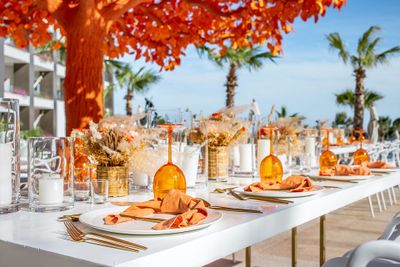 Elegant outdoor dining setup with orange napkins and glassware on a white table, surrounded by palm trees and a vibrant orange-leaved tree.