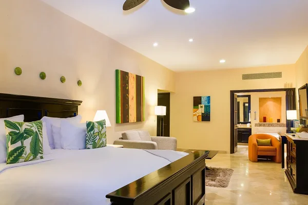 Spacious hotel room with a large bed, tropical-themed pillows, armchair, and modern art. Open doorway leads to a well-lit bathroom.