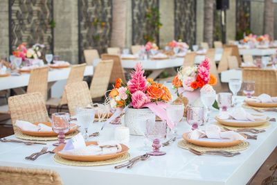 Elegant outdoor dining setup with white tables, wicker chairs, and vibrant floral centerpieces. Plates and glasses are neatly arranged.