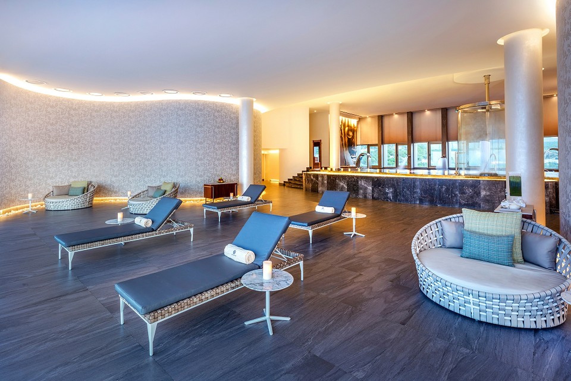 Spacious, modern spa lounge with sleek loungers, round seating, ambient lighting, and large windows overlooking greenery.