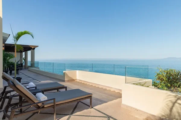 Oceanfront terrace with two lounge chairs, glass railing, and a clear blue sky. Palm plant and distant mountains enhance the serene view.